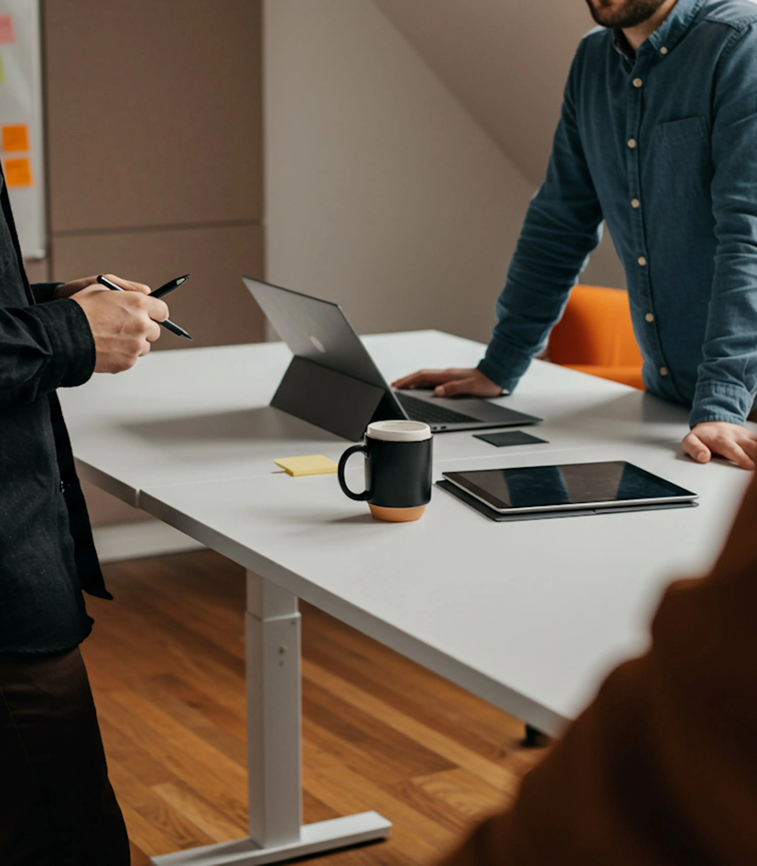Colleagues collaborating around a white office table with a laptop, tablet, and coffee mug in a modern workspace.