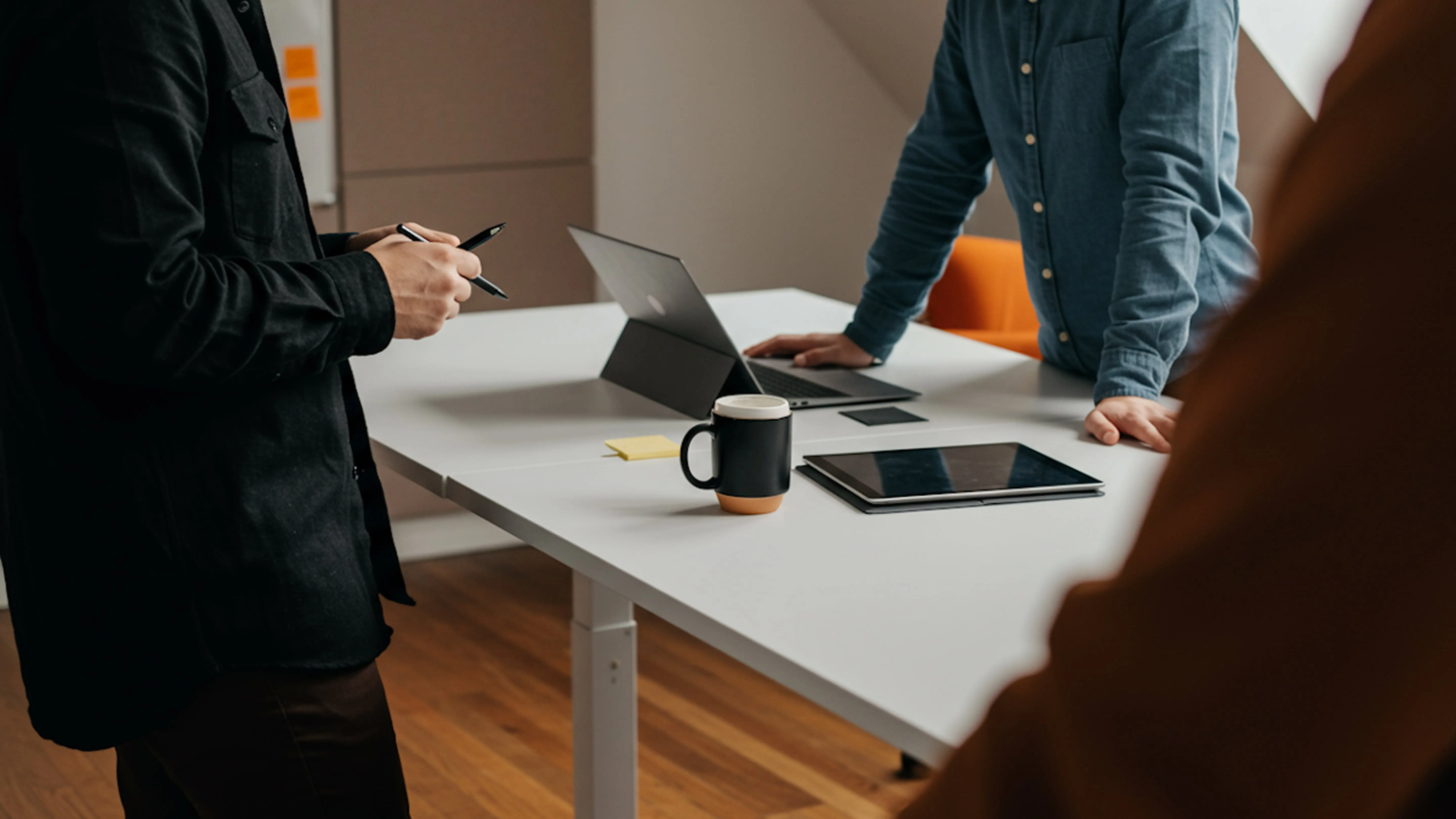 Colleagues collaborating around a white office table with a laptop, tablet, and coffee mug in a modern workspace.