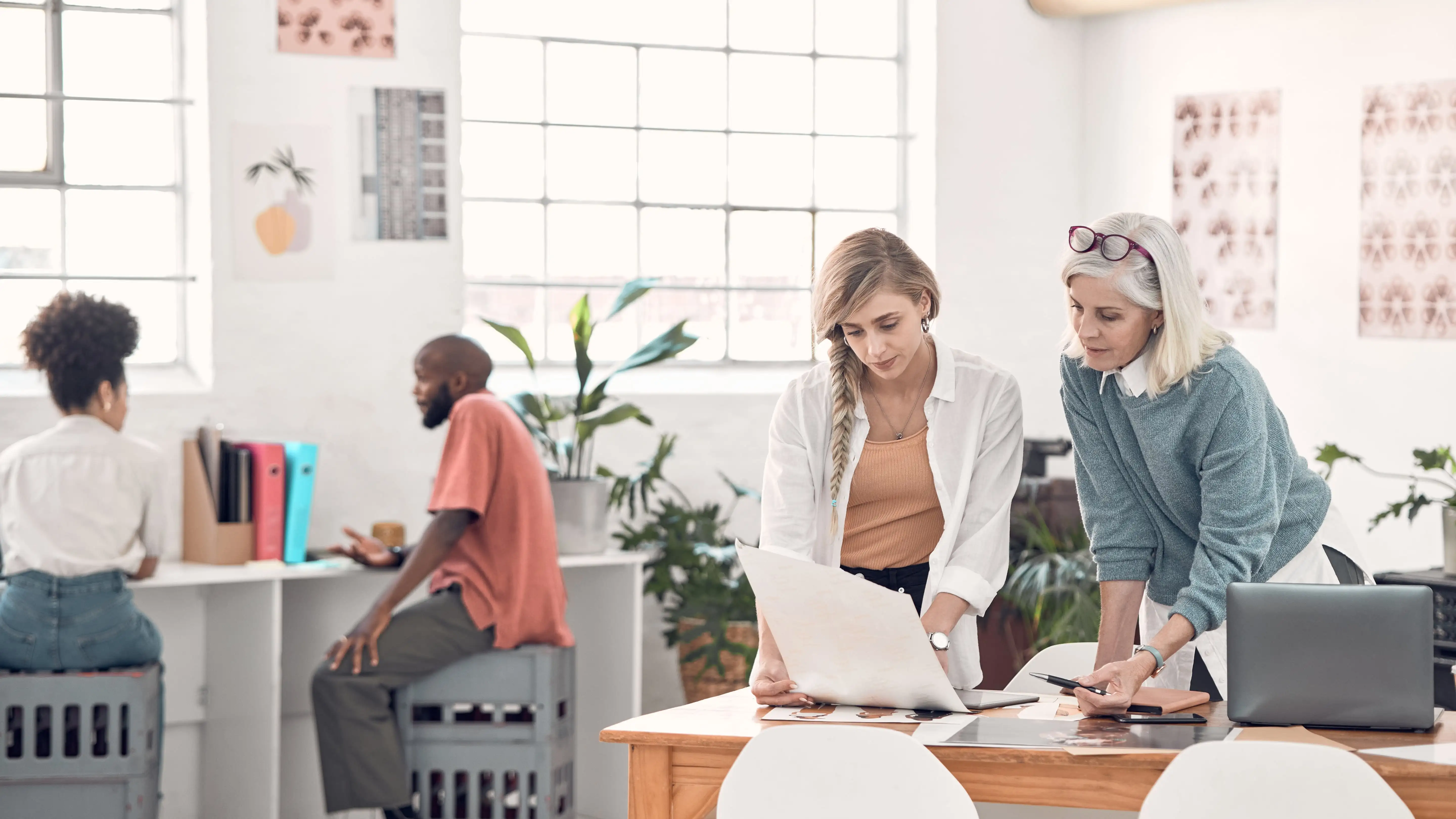 Four people working in a modern, open-plan office; two women are consulting over documents in the foreground.